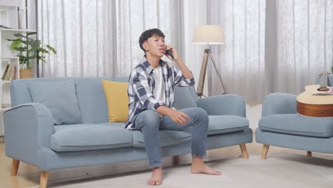 full body of asian teen boy talking on smartphone while sitting on sofa in the living room at home