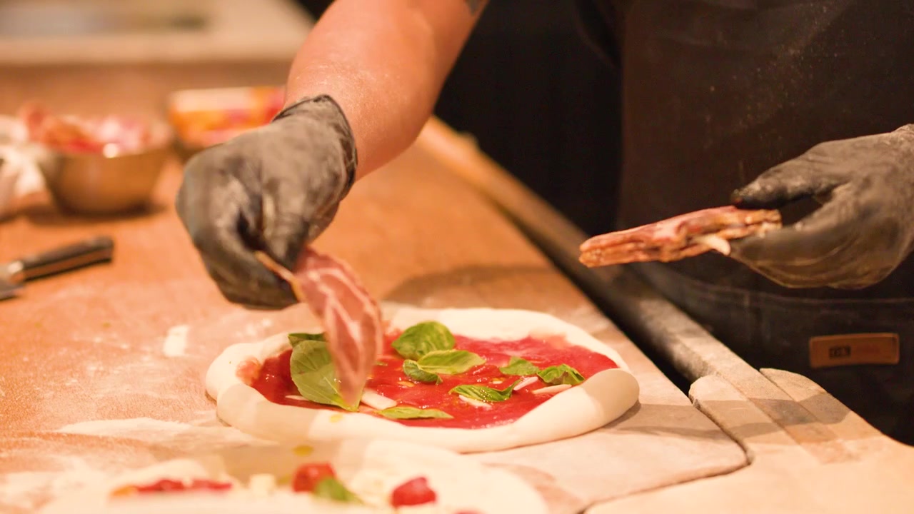 Primo piano delle mani dello chef che posizionano il prosciutto su impasto di pizza crudo con salsa di pomodoro e basilico.