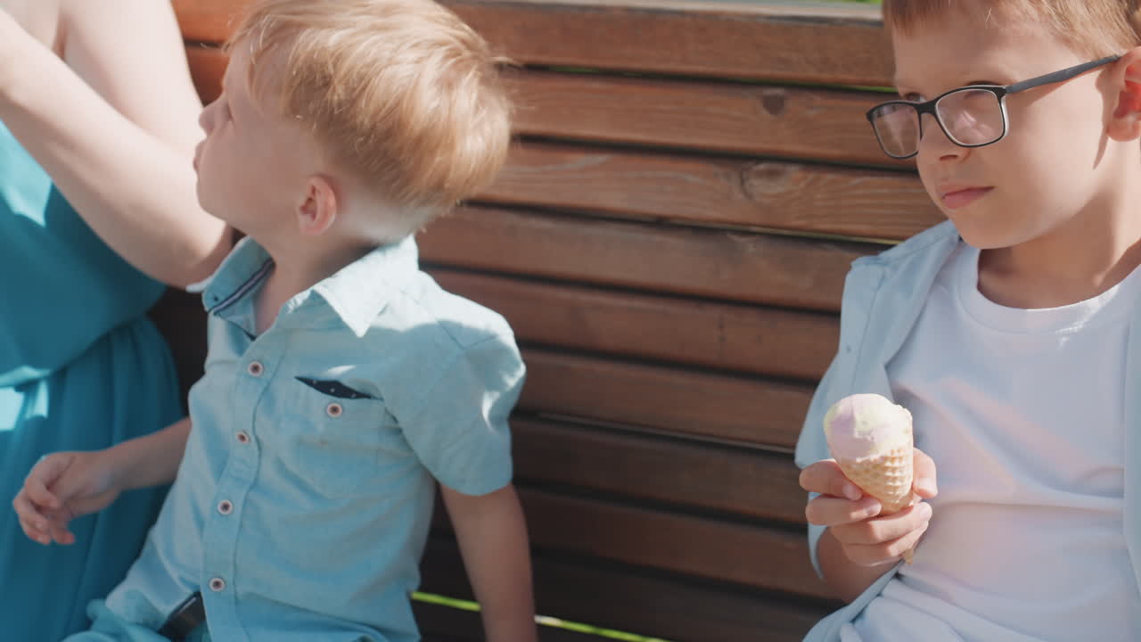 Ragazzo seduto su una panchina di legno con un gelato, sguardo abbassato dietro gli occhiali, espressione pensierosa, un fratello/sorella minore accanto, sentiero del parco illuminato dal sole, atmosfera tranquilla di un fine settimana in famiglia, momento spontaneo e contemplativo.