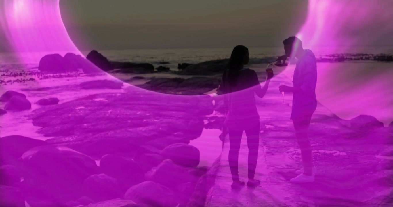 Coppia che brinda in spiaggia mentre un anello di animazione tecnologica emerge e si espande sull'acqua.