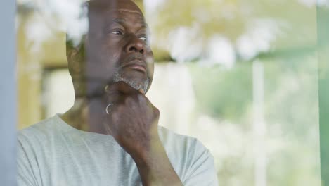 Thoughtful-african-american-senior-man-standing-in-living-room-holding-chin,-looking-out-of-window