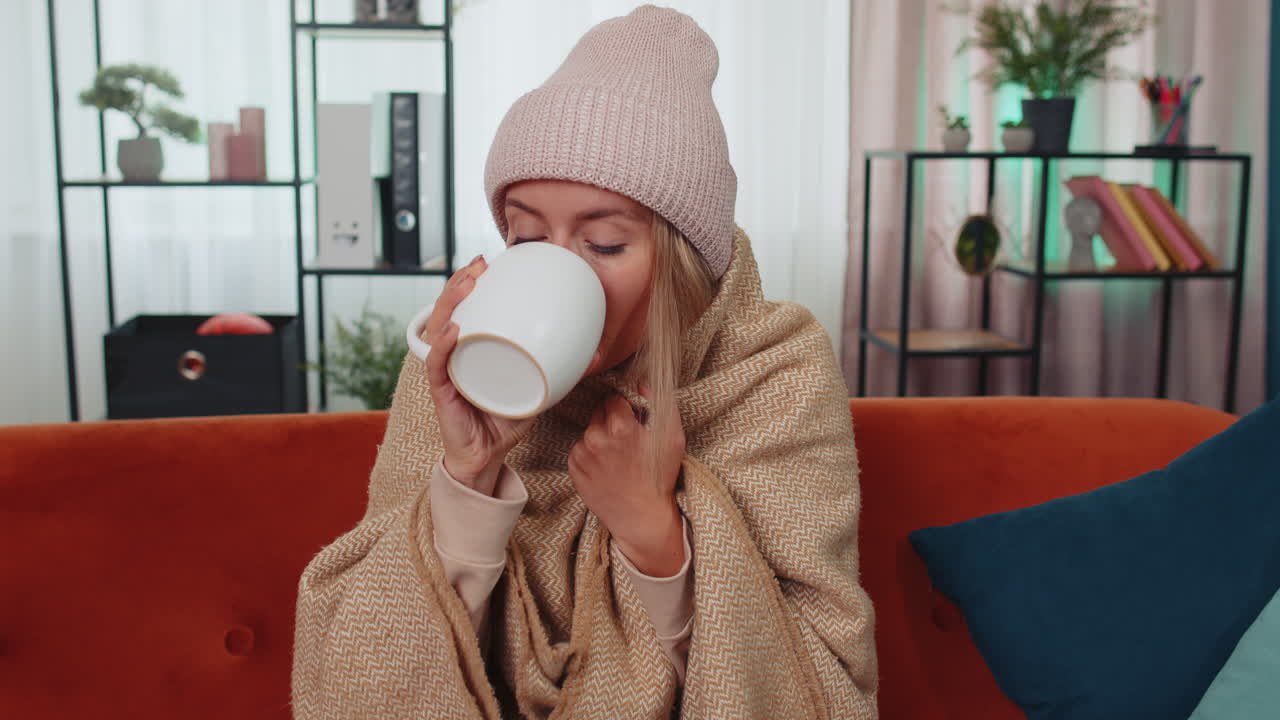 Una giovane donna malata indossa un cappello avvolto in un quadri si siede da sola tremante dal freddo sul divano bevendo tè caldo