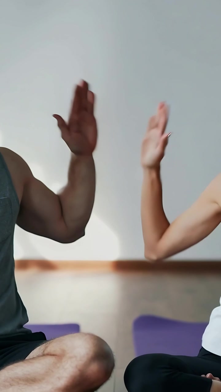 Video vertical: Pareja en camisetas de tirantes soltando las manos y levantando los brazos para chocar los cinco en casa, sobre colchonetas moradas.