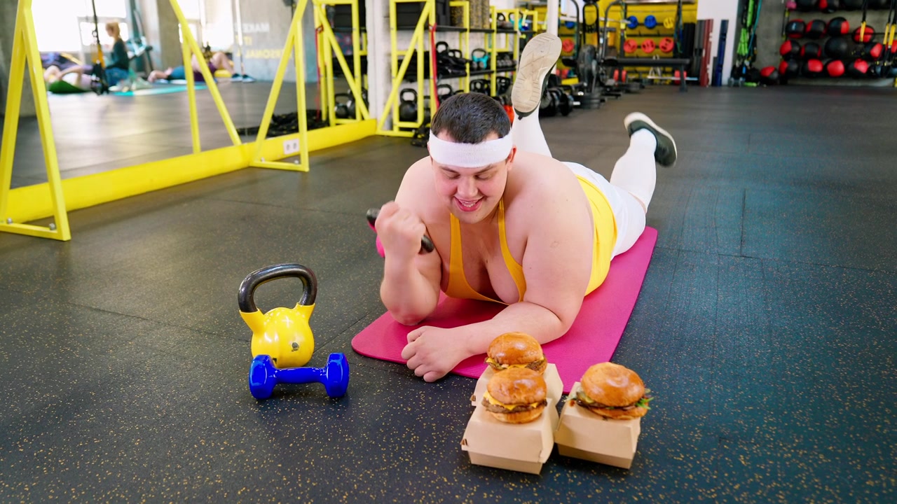 Uomo in sovrappeso divertente che si allena con un manubrio accanto a degli hamburger