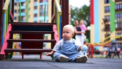 sweet baby boy with blond hair sits down at the wooden stairs in the playground. cute toddler holds a water bottle in hands. blurred backdrop.