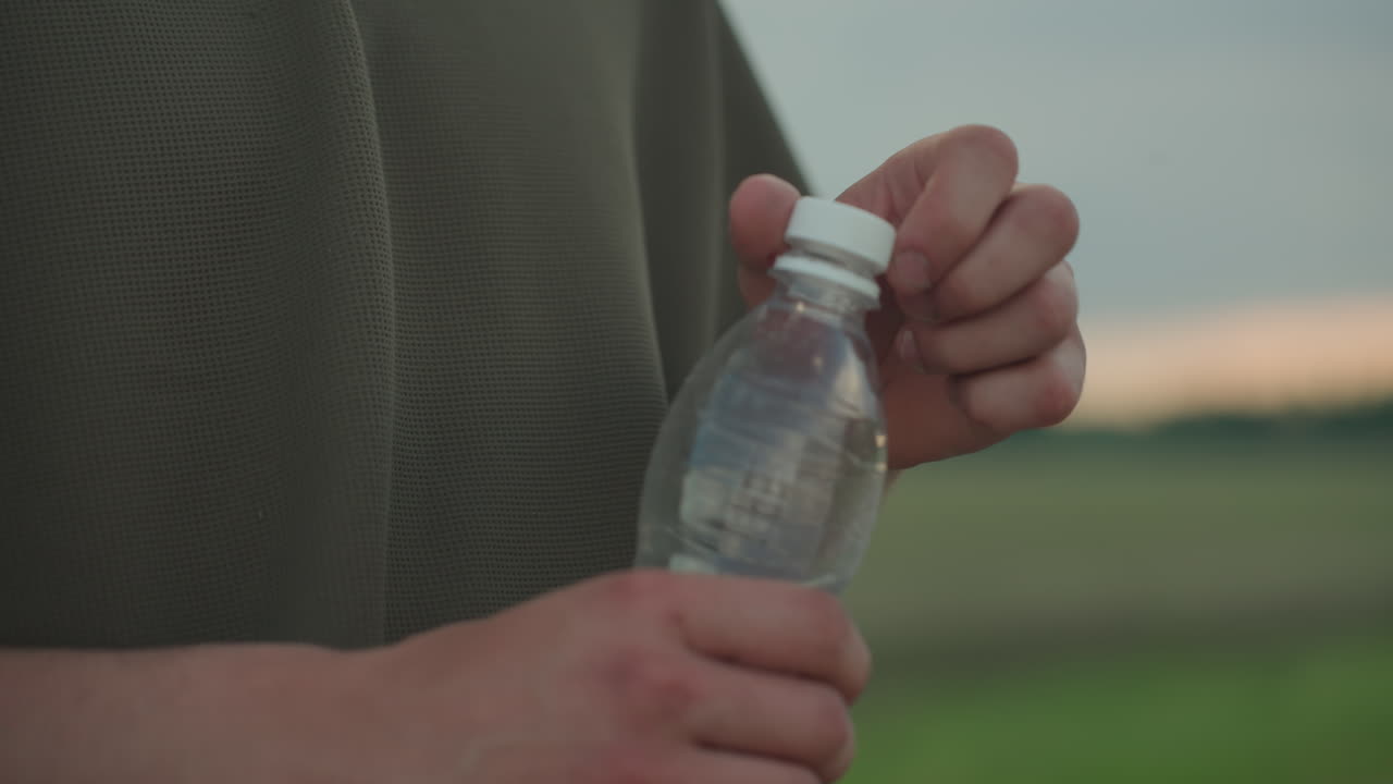 close up of man hands tightening cap on clear plastic water bottle outdoors with green blurred background, focus on tactile motion and hydration gesture