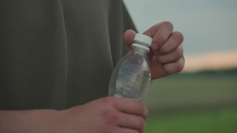 close up of man hands tightening cap on clear plastic water bottle outdoors with green blurred background, focus on tactile motion and hydration gesture