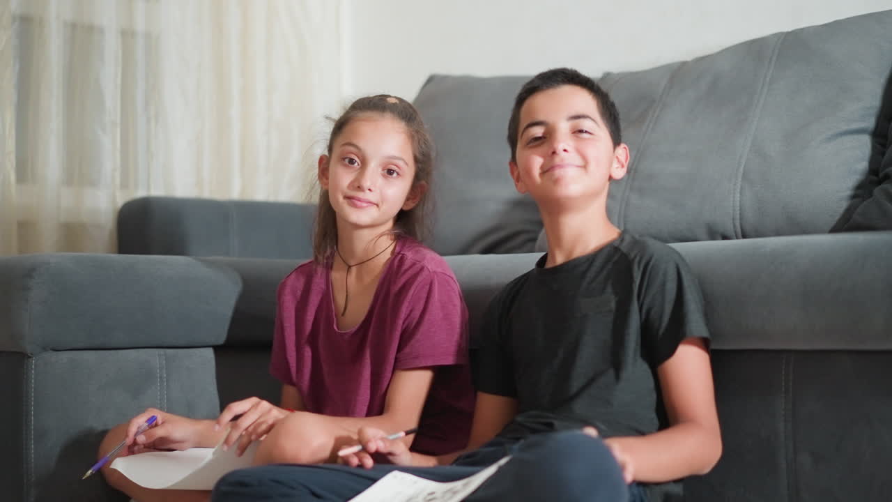 Primos adorables sentados en el suelo cerca del sofá, sujetando un papel y sonriendo con calidez a la cámara en un ambiente interior acogedor, mostrando la alegría de la actividad creativa compartida y el tiempo de unión con expresiones amistosas y relajadas