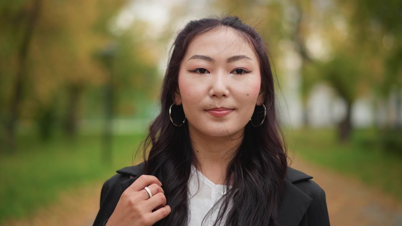 Primer plano de una mujer joven con cabello largo y oscuro, usando grandes aretes de aro y sujetando la solapa de su abrigo negro en un parque al aire libre. La escena presenta luz natural suave y áreas de césped verde.