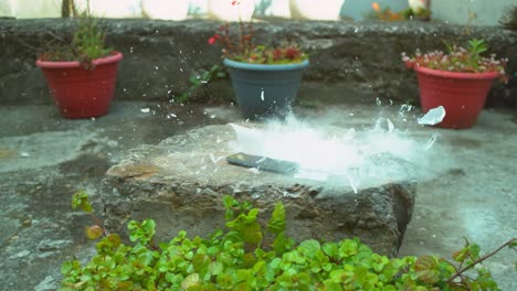 Glass-cup-falls-onto-a-phone-in-a-garden,-shattering-and-emitting-smoke-in-motion