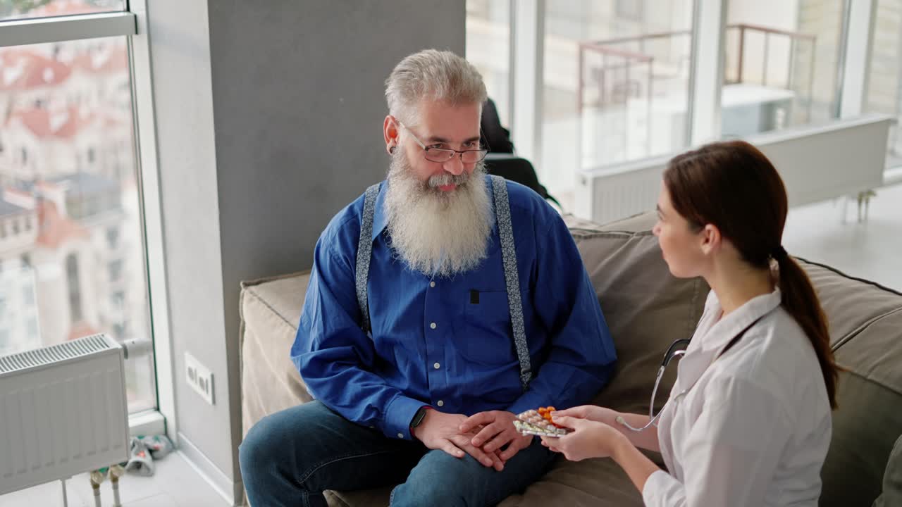 A happy elderly man with gray hair and a lush beard in glasses in a blue shirt takes pills in his hands that were given to him by a brunette doctor girl in a medical gown sitting on a brown sofa in a modern apartment