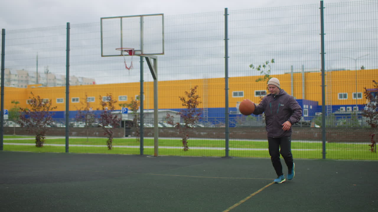 Campo da basket all'aperto: uomo che palleggia sotto un cielo nuvoloso, mentre si esercita da solo nel riscaldamento e negli esercizi di tiro accanto a un parco recintato con un edificio industriale giallo sullo sfondo; il berretto e la giacca suggeriscono