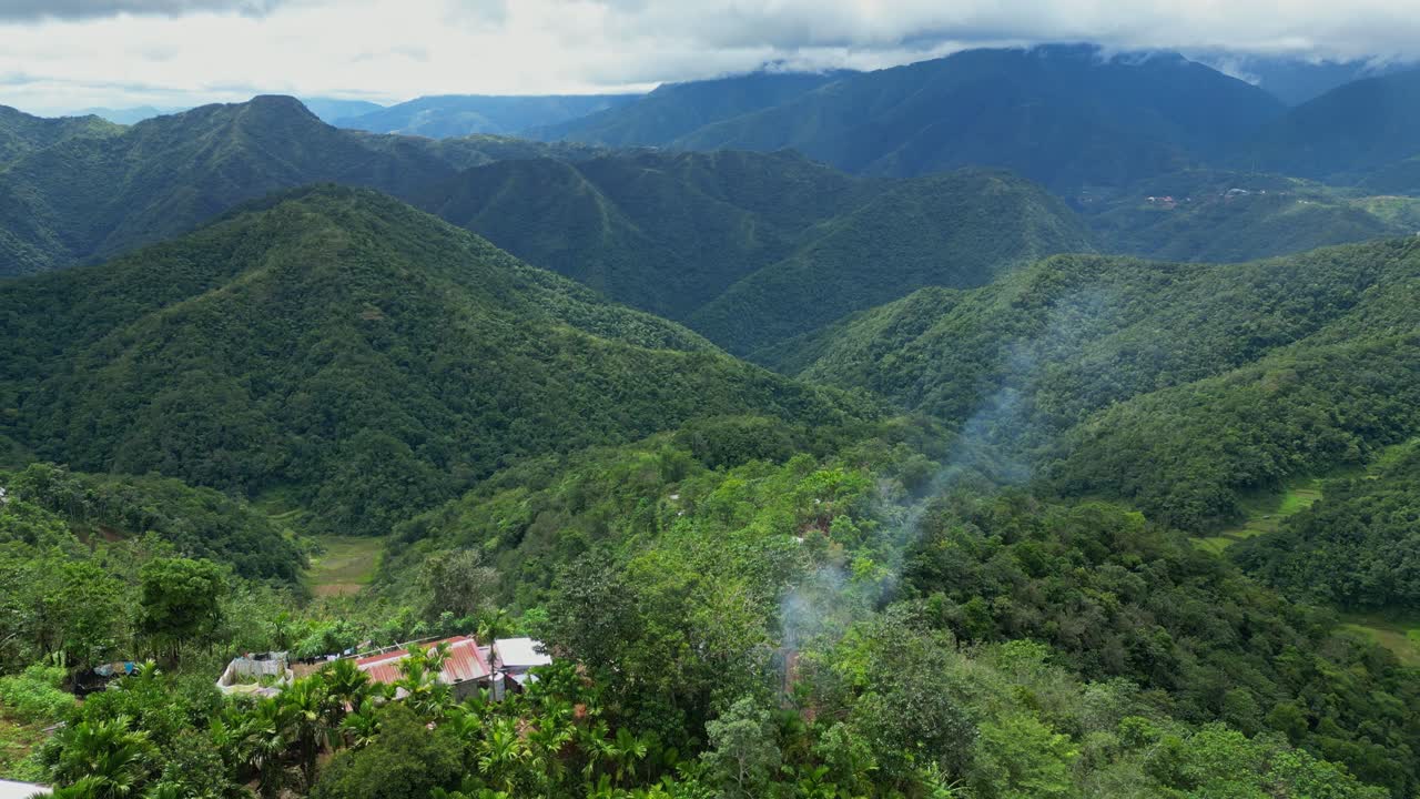 Hướng lên trên không trung vượt qua những ngôi nhà vùng núi nông thôn ở Banaue, Ifugao, lướt về phía làn khói bốc lên với những dãy núi rộng lớn, xếp lớp ở phía xa tại Banaue, Ifugao, Philippines