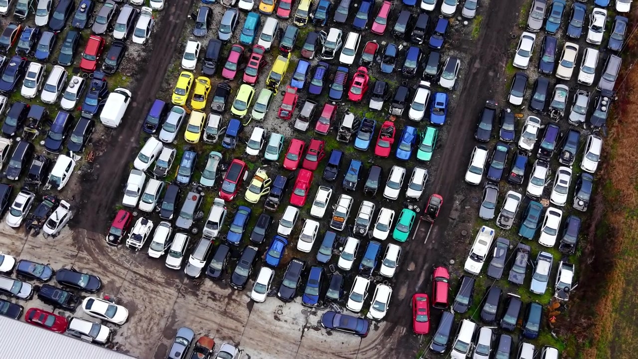 Veduta dall'alto di un deposito di veicoli rottamati. File fitte di auto danneggiate bianche, blu e rosse viste direttamente dall'alto di un cortile sterrato.