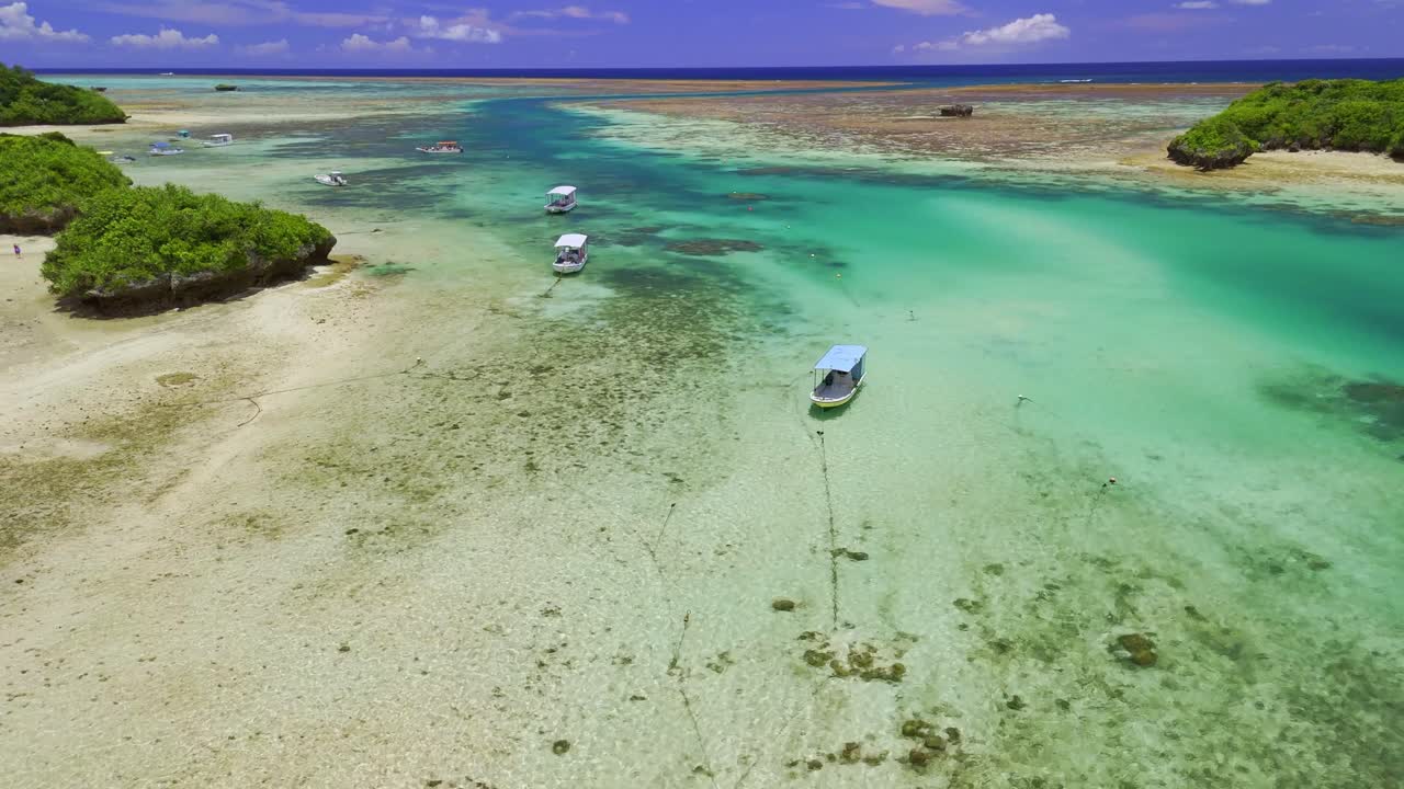 A stunning bird's-eye view tracking a lone boat as it moves through the crystal-clear, shallow turquoise waters of a tropical lagoon, with the sandy seabed clearly visible below
