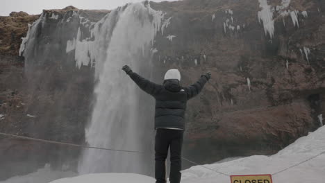 Parte-Posterior-Del-Hombre-En-Ropa-De-Invierno-Abrigada-Caminando-Sobre-La-Nieve-Bajo-Una-Cascada-En-Islandia