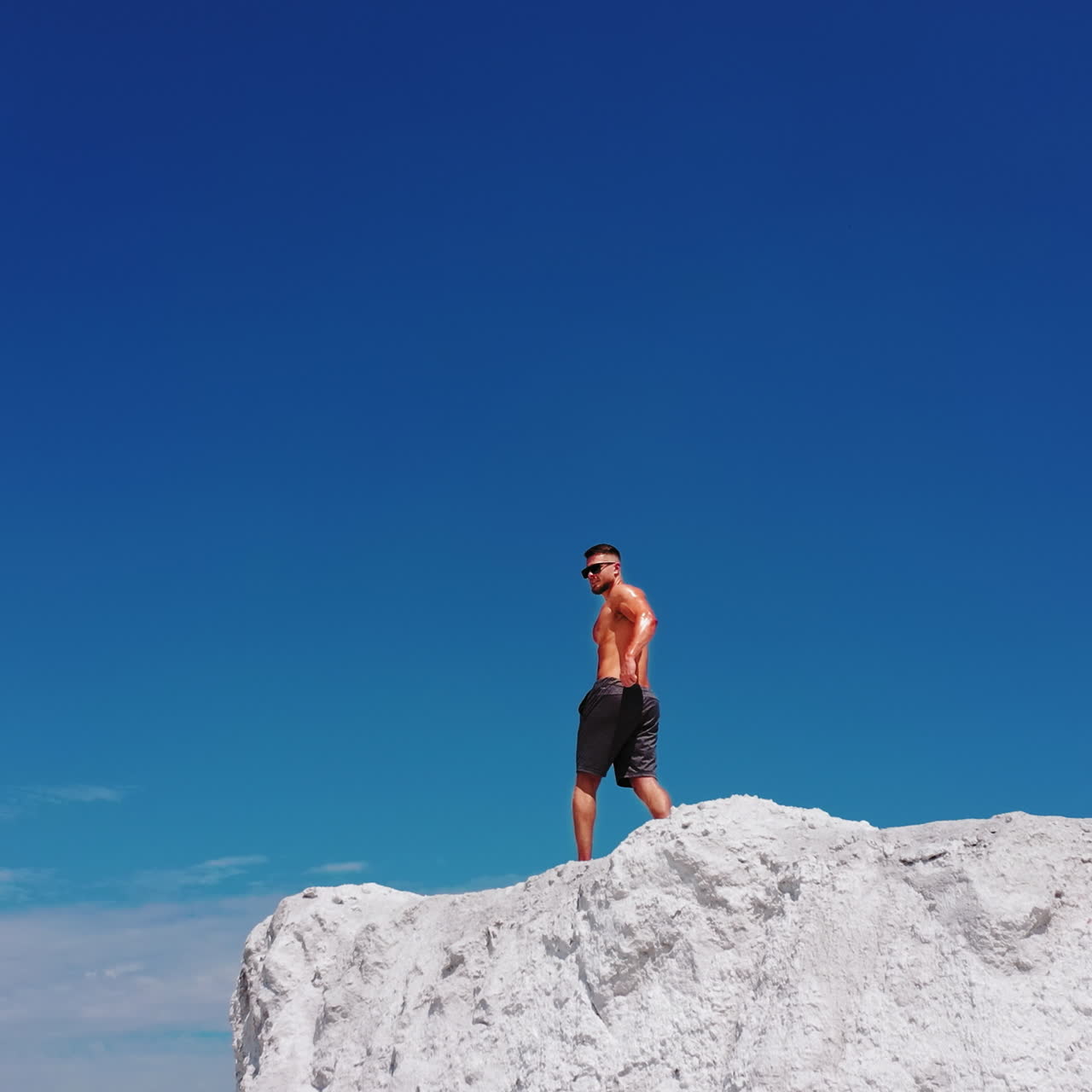 Uomo muscoloso su collina bianca. Bodybuilder senza maglietta in posa sulla cima della montagna sotto il cielo blu. Telecamera in movimento all'indietro
