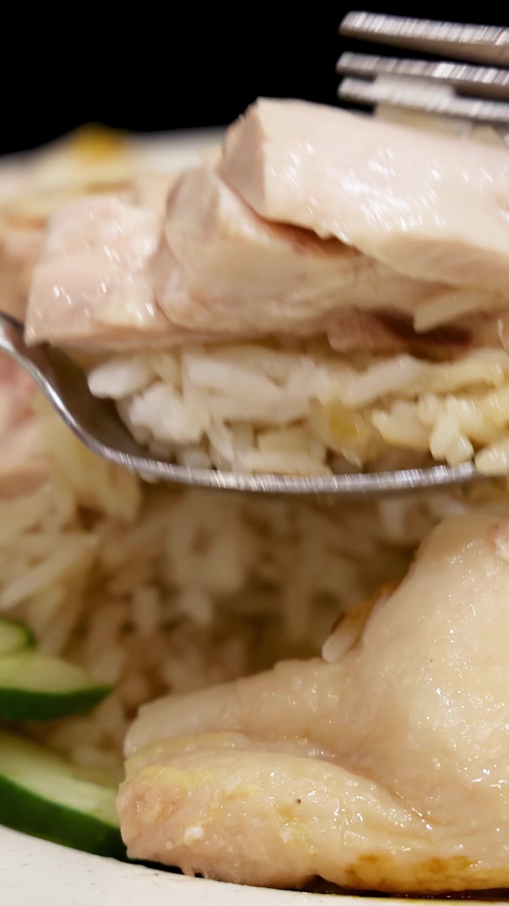 Primo piano di un cucchiaio e una forchetta che raccolgono tenero pollo Hainanese con riso da un piatto sotto un'illuminazione brillante e uniforme. Movimento minimo della telecamera