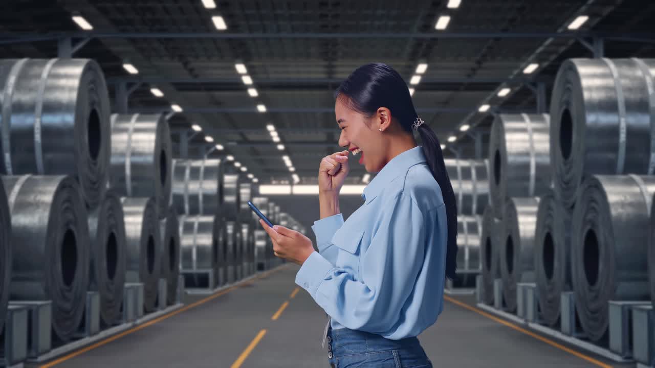 Vista lateral de uma trabalhadora profissional asiática com seu smartphone, perto de pilhas de grandes bobinas de aço em um armazém, levantando o punho em comemoração após verificar o smartphone.