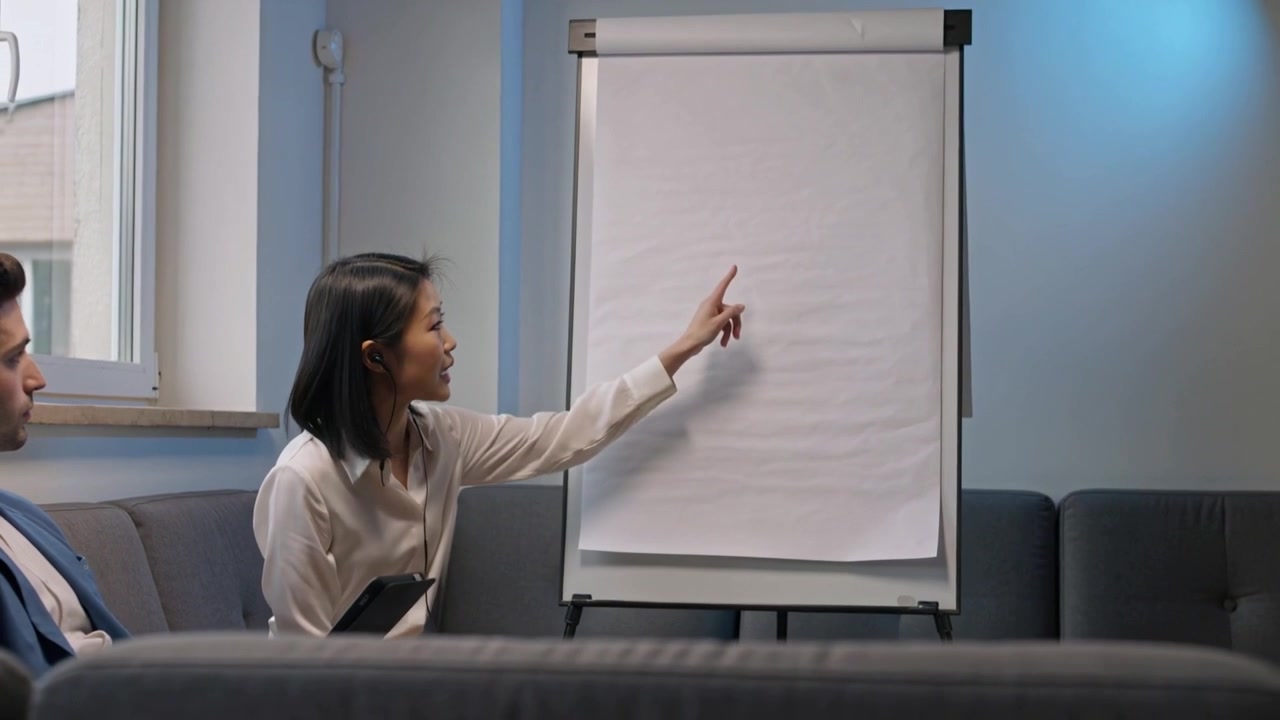 Una donna in primo piano con top bianco e tablet indica un flipchart all'inizio di una riunione in una lounge.
