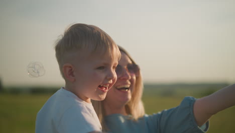 A cheerful woman in a blue dress holds a little boy in a white shirt, both laughing joyfully as they reach out to touch colorful bubbles floating around them in a sunny grassy field