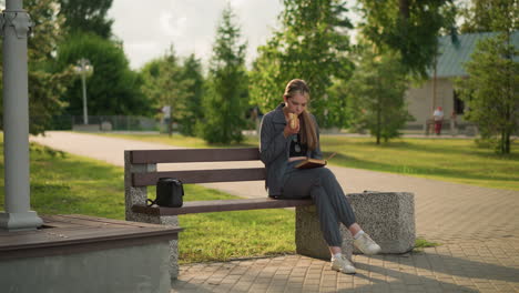 mujer sentada en un banco del parque sosteniendo un bocadillo y un libro, comiendo y leyendo con una bolsa negra a su lado, en un entorno al aire libre pacífico rodeado de árboles y vegetación, fondo borroso con personas