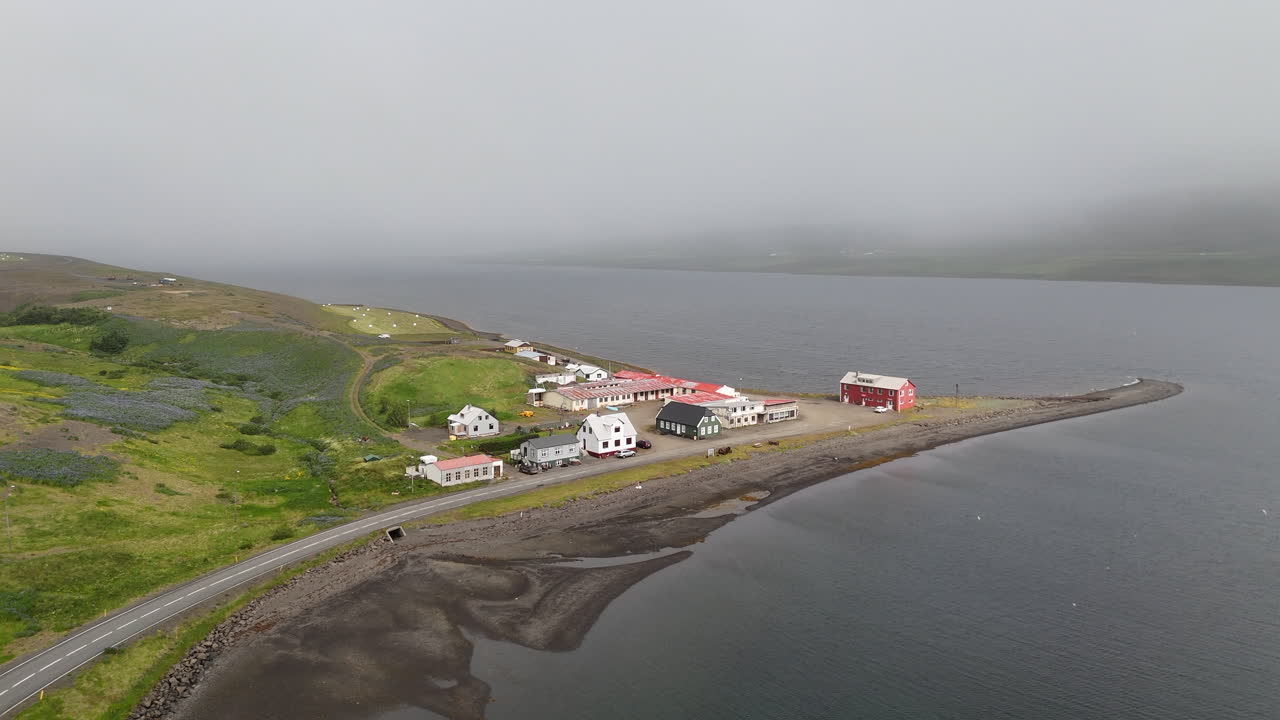 Small coastal village of Borðeyri with its curved shoreline and scattered houses, seen under low-hanging misty clouds drifting over the fjord
