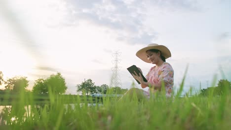 Asiatische-Frau-Benutzt-Ein-Tragbares-Gerät,-Das-Auf-Einer-Wiese-In-Der-Nähe-Des-Flusses-Sitzt