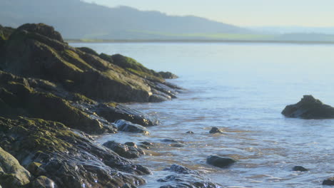 Sea-gently-breaking-against-rocks-on-shoreline-with-misty-hills-in-distance