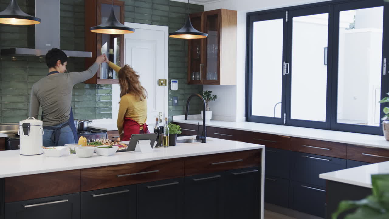 Gelukkig Kaukasisch lesbisch stel dat eten bereidt, danst en een tablet gebruikt in een zonnige keuken, met ruimte voor tekst