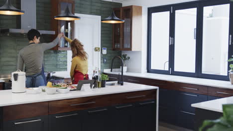 Happy-caucasian-lesbian-couple-preparing-food,-dancing-and-using-tablet-in-sunny-kitchen,-copy-space