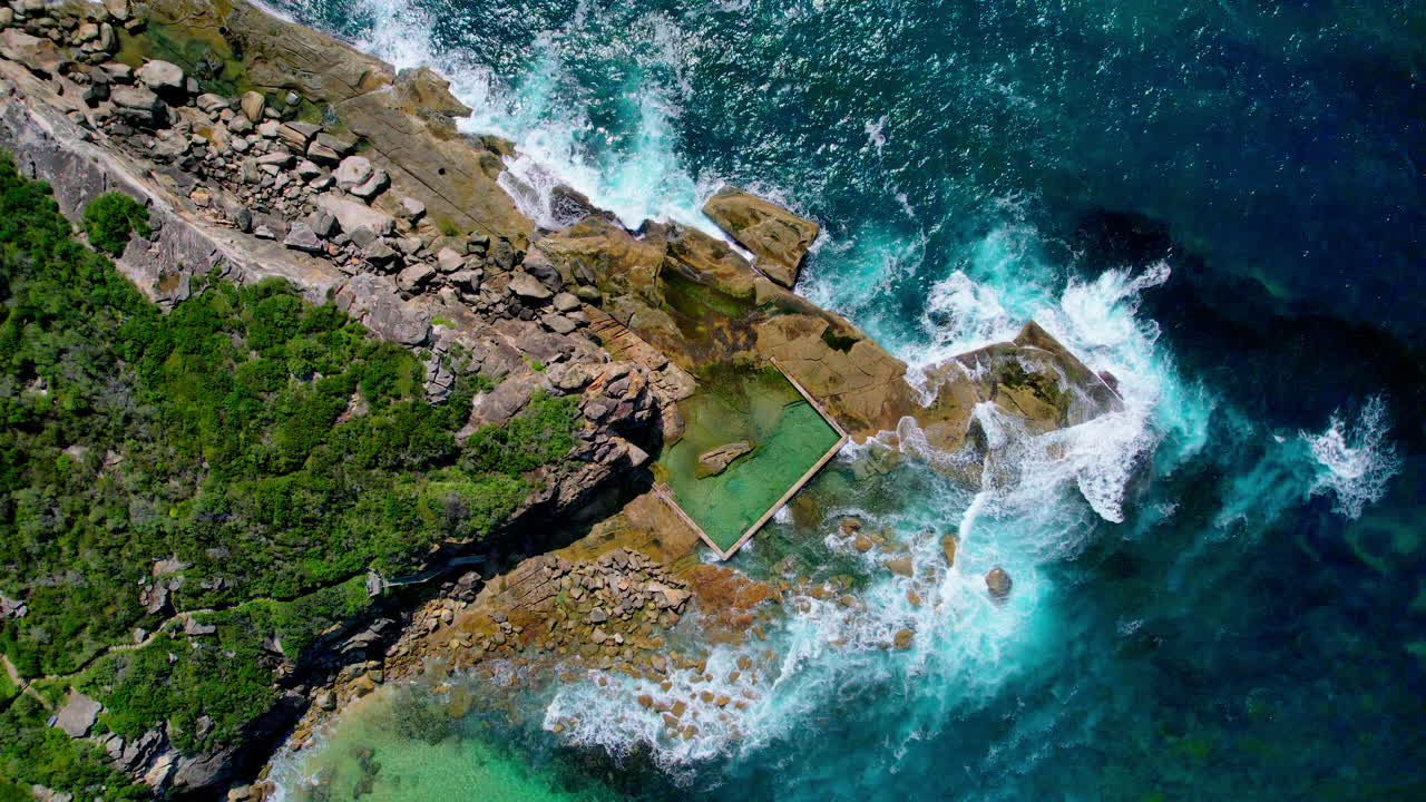 Aerial top view of North Curl Curl Rock Pool in Sydney. Clear turquoise water, waves crashing nearby, and rocky coastline create a stunning, natural coastal scene from above.