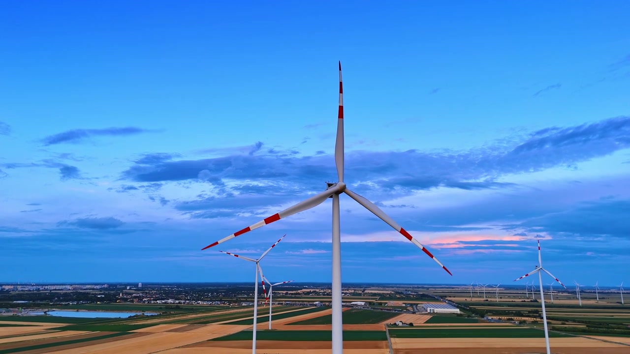 Turbina eolica industriale sotto un drammatico cielo crepuscolare blu. Inquadratura dal basso di un'acciaieria bianca all'orizzonte con una debole luce del tramonto sulle nuvole.