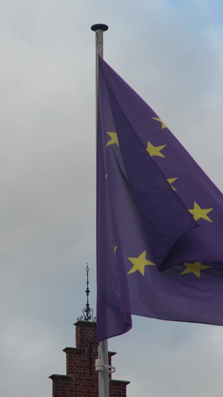Bandiera dell'Unione Europea sventola vigorosamente in cima a un edificio storico contro un cielo nuvoloso, catturata con telecamera stabile.