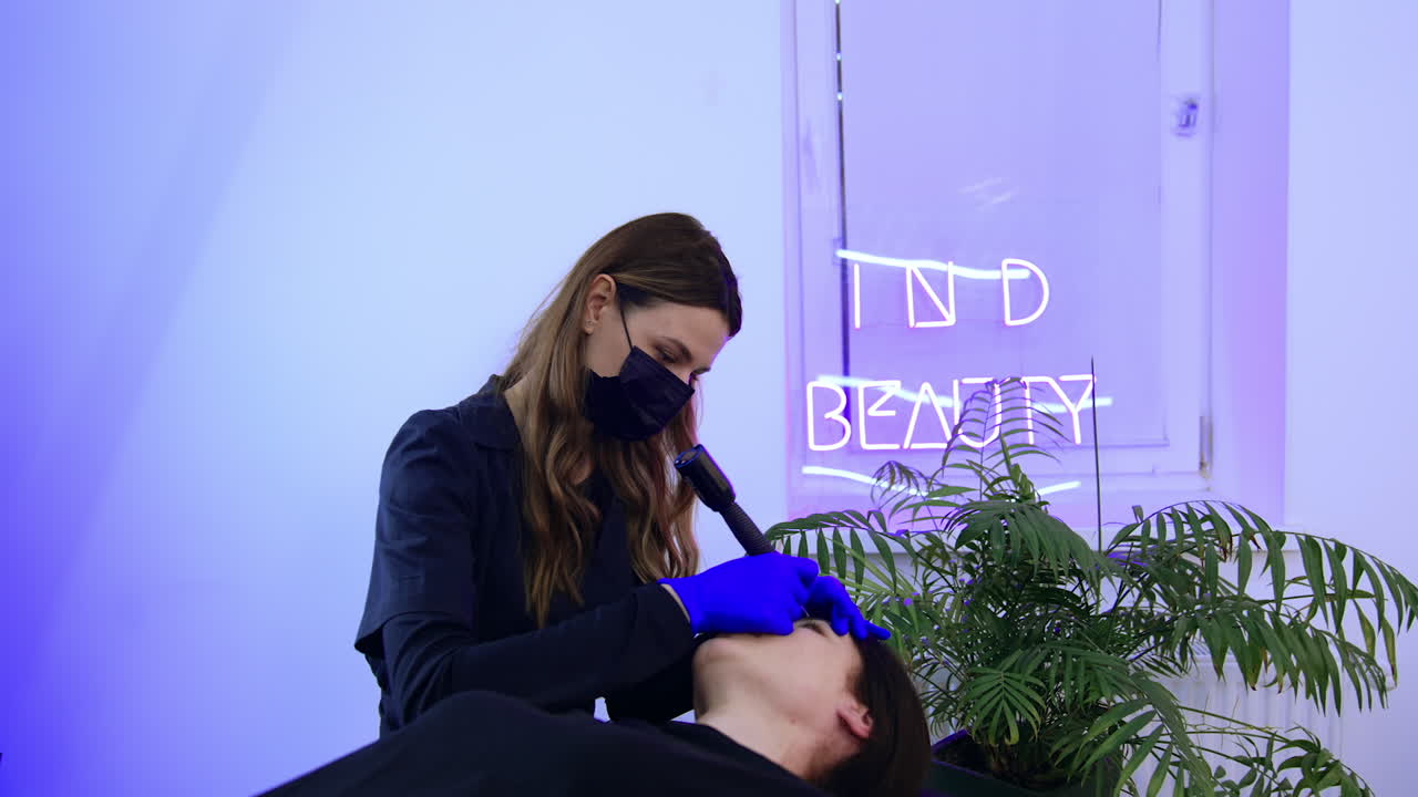 Caucasian brunette female lies on the couch in beauty salon. Beautician carrying out the procedure of permanent make up for eyebrows.
