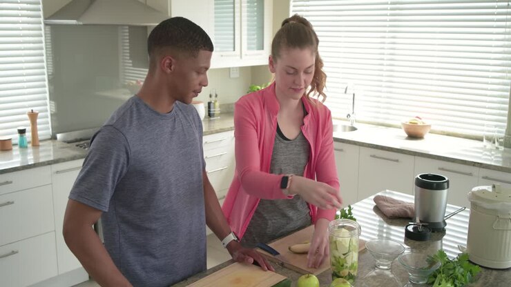Couple preparing smoothie man pouring blue pitcher into jar while woman slicing banana in kitchen