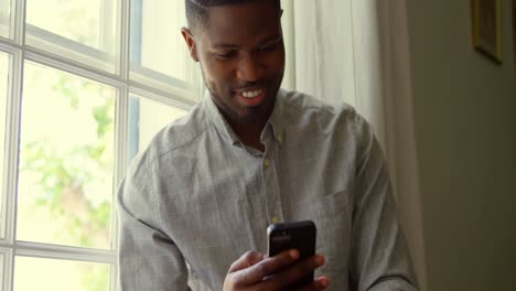 Front-view-of-young-black-man-using-smartphone-and-sitting-on-window-sill-in-a-comfortable-home-4k
