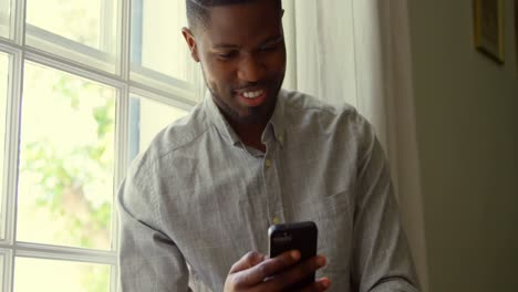 front view of young black man using smartphone and sitting on window sill in a comfortable home 4k