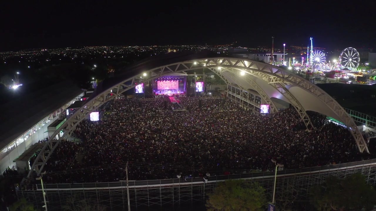 Nighttime aerial drone view of a massive crowded outdoor concert venue under a huge arched steel roof structure, stage with glowing pink LED screens and bright concert lights and carnival games.