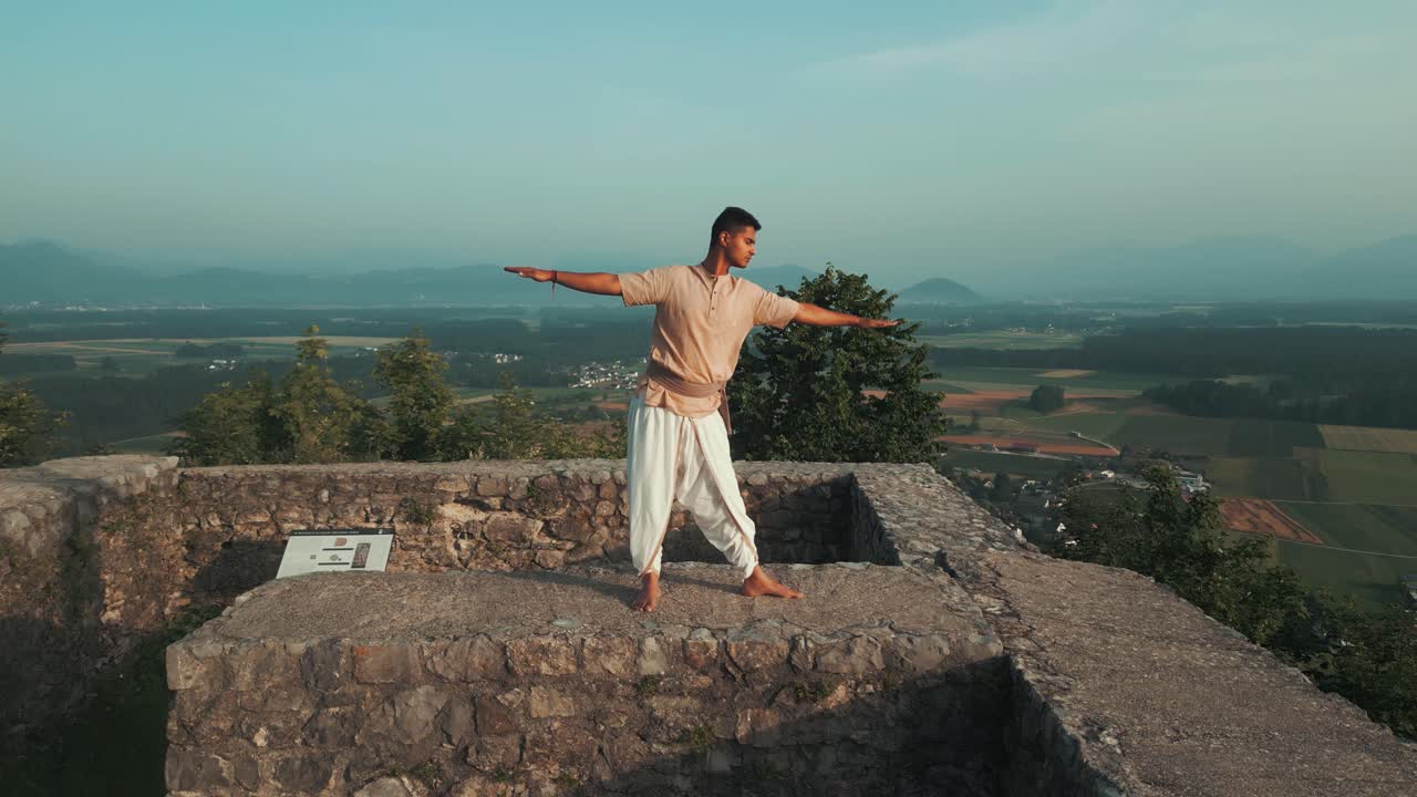 hombre indio haciendo postura de hatha yoga al amanecer en la pared del castillo de piedra en la cima de la colina descalzo y con ropa tradicional de meditación con campos y árboles detrás de él