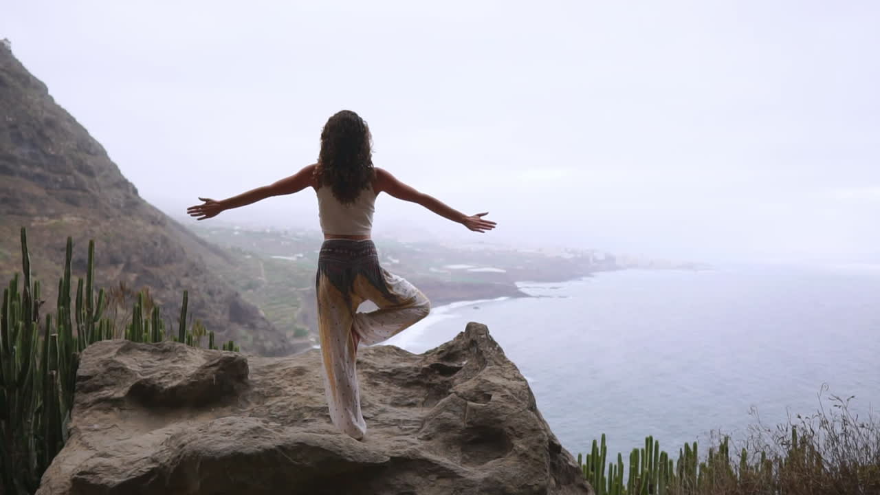 In slow motion, woman practices yoga's warrior pose by ocean, beach, and rocky mountains, symbolizing motivation, inspiration, and unity of fitness and nature in a healthy lifestyle