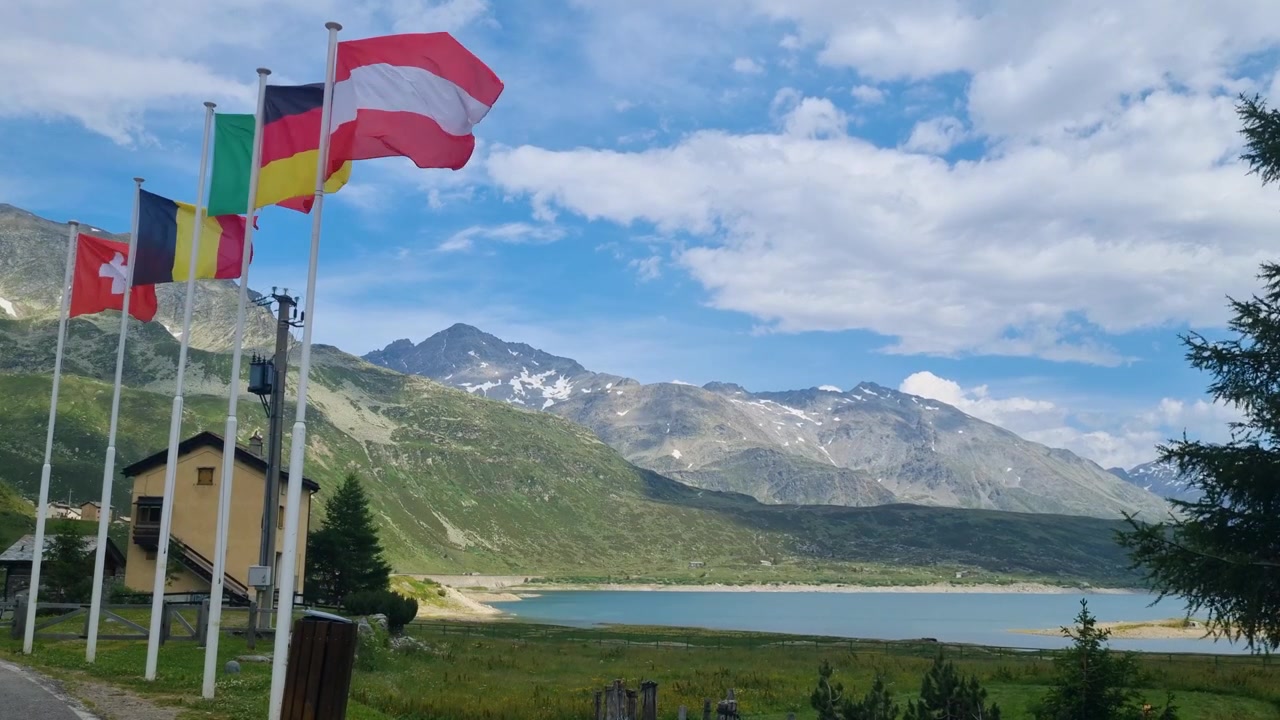 Vlajky Rakouska, Německa, Itálie, Belgie a Švýcarska vlají nad jezerem Lago di Montespluga za oblačného dne, symbolizující mezinárodní cestování v Alpách a přeshraničního ducha evropských hor.