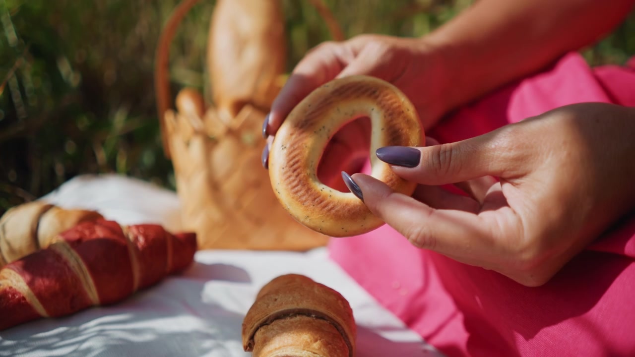 Woman with dark nail polish reaches for and holds croissant among various baked goods on white cloth outdoors. scene includes bagels red croissants soft lighting creating shadows from tree