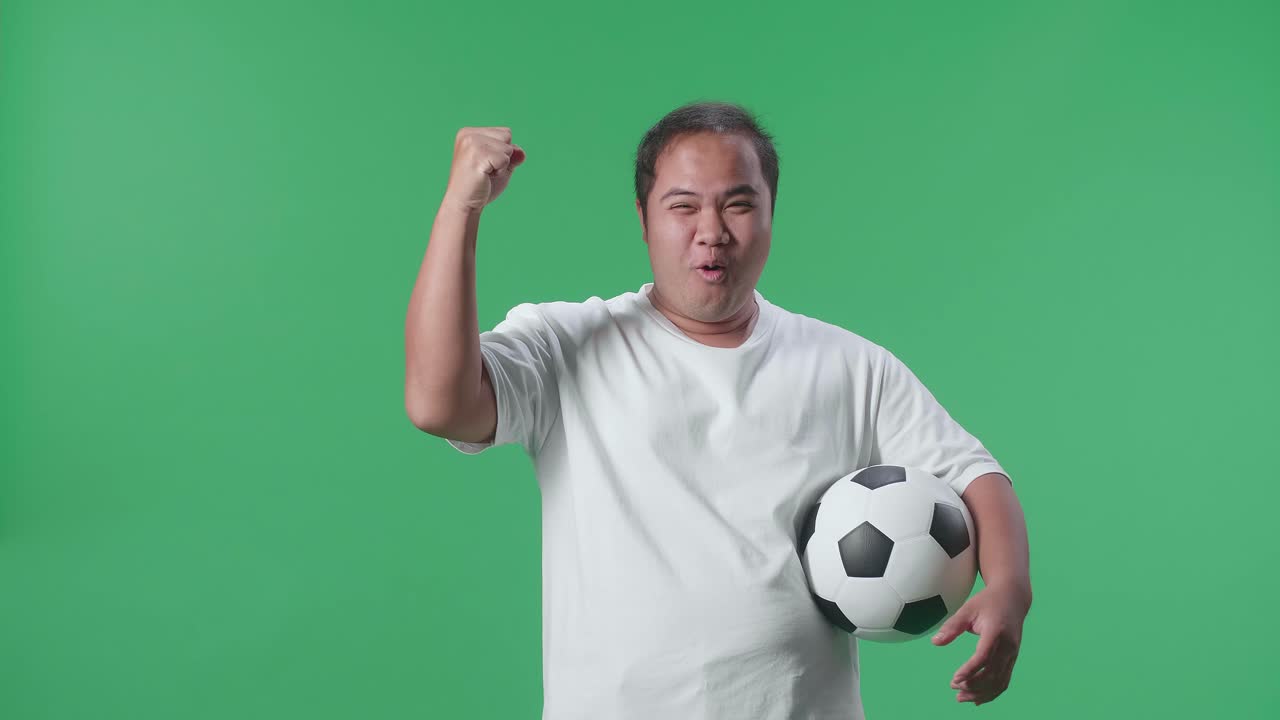 공을 들고 있는 아시아인 남자가 골을 외치며 우승팀을 축하하고 초록색 스크린 배경에서 축구를 응원하고 있다.