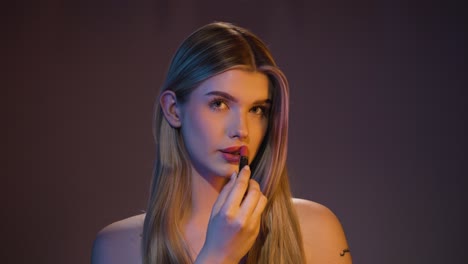 medium slow motion shot of a young attractive woman putting red lipstick on her lips and giving a lewd look to the camera in front of black and brown background