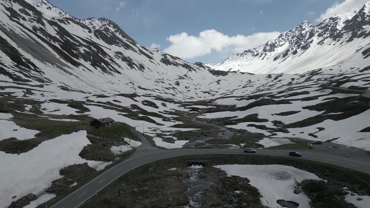 Drone raising backward over a snowy alpine valley, capturing two cars driving along a winding mountain road.
