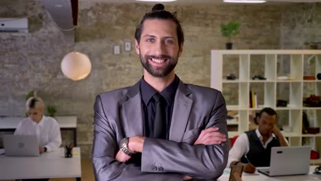 happy businessman with beard is standing with arms crossed and watching at camera in office, smiling, colleagues are working with laptops, work concept