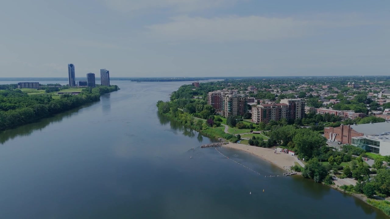 Vista aerea del lungofiume cittadino con edifici alti e vegetazione. L'acqua calma riflette il cielo e le strutture distanti. Il paesaggio urbano si fonde con la natura in una tranquilla luce diurna. Perfetto per paesaggi urbani