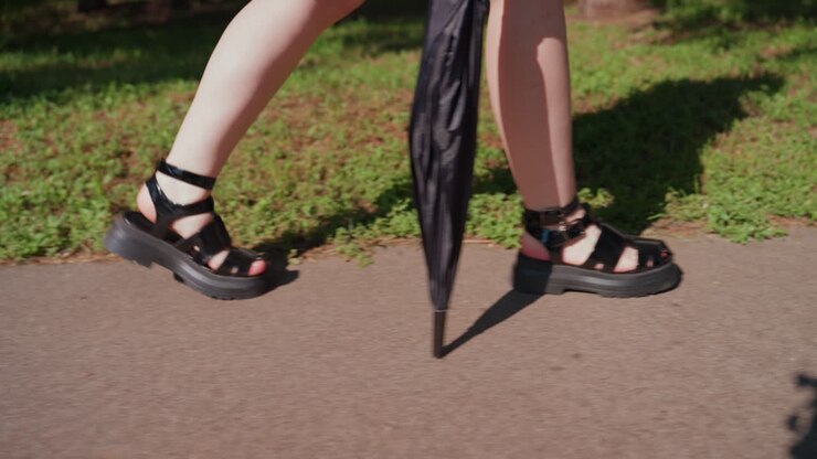gentle woman ambles through park, serene lady moves slowly on shaded park walkway with umbrella, tranquil woman with folded umbrella and sandals reflecting gentle sunlight on peaceful park trail