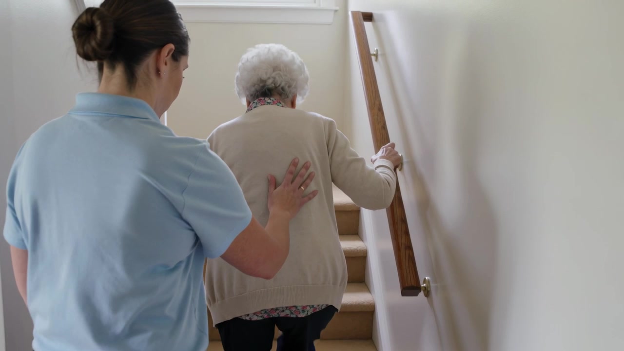L'assistente, con una mano dietro di lei, la guida su per le scale di casa fino al pianerottolo, tenendo il corrimano e indossando una polo.